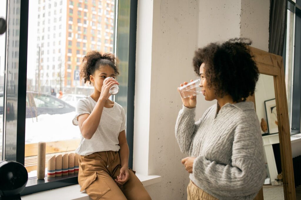 Positive African American mother with daughter in casual wear looking at each other while drinking water from glasses in light room
